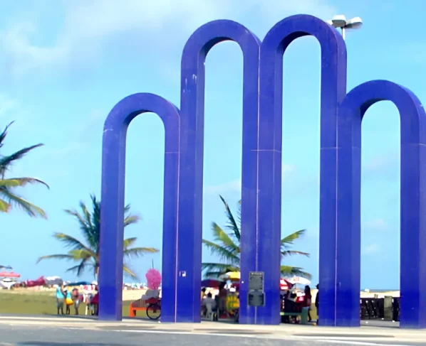 Monumento dos Arcos da Orla de Atalaia, em Aracaju, com coqueiros e praia ao fundo.