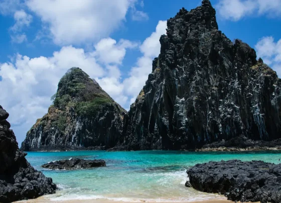 A imagem mostra uma paisagem costeira tropical de origem vulcânica, com formações rochosas escuras emergindo do mar e águas cristalinas em tons de azul-turquesa.