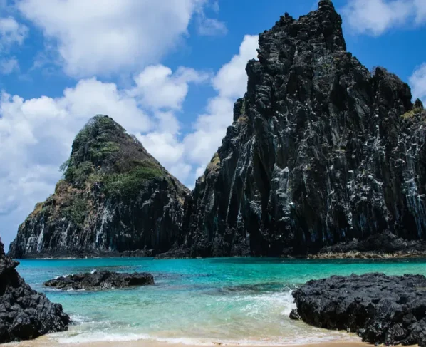 A imagem mostra uma paisagem costeira tropical de origem vulcânica, com formações rochosas escuras emergindo do mar e águas cristalinas em tons de azul-turquesa.