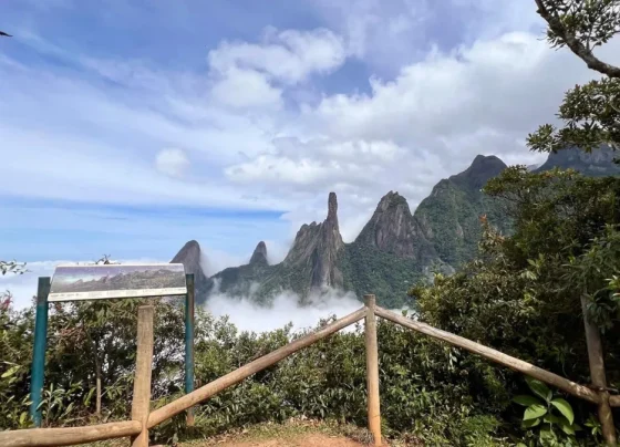 Vista panorâmica do Dedo de Deus, na Serra dos Órgãos, acima de um mar de nuvens, vista a partir de um mirante com cerca de madeira e placa informativa.
