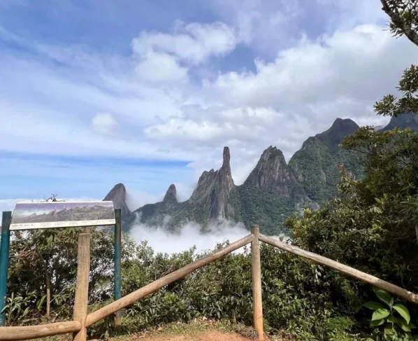 Vista panorâmica do Dedo de Deus, na Serra dos Órgãos, acima de um mar de nuvens, vista a partir de um mirante com cerca de madeira e placa informativa.