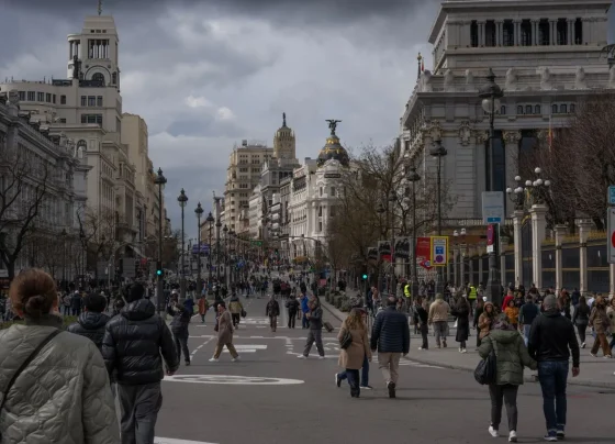 Vista da avenida Gran Vía em Madri repleta de pedestres caminhando, com edifícios históricos imponentes e o famoso Edifício Metrópolis ao fundo sob céu nublado.