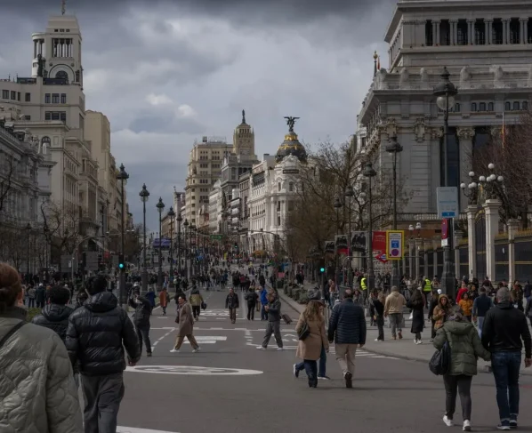 Vista da avenida Gran Vía em Madri repleta de pedestres caminhando, com edifícios históricos imponentes e o famoso Edifício Metrópolis ao fundo sob céu nublado.