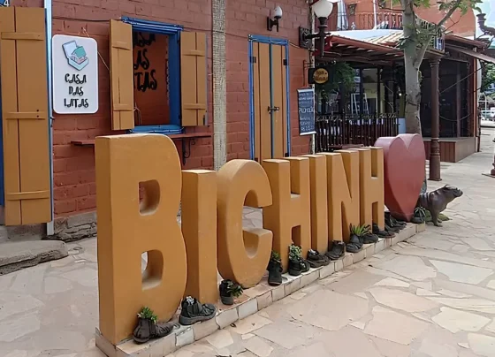 Escultura grande com as letras "BICHINHO" em um tom de amarelo-ocre, posicionada em uma calçada de pedra em frente à loja "Casa das Latas", com sapatos antigos servindo como vasos de plantas ao pé das letras e um coração vermelho ao lado.