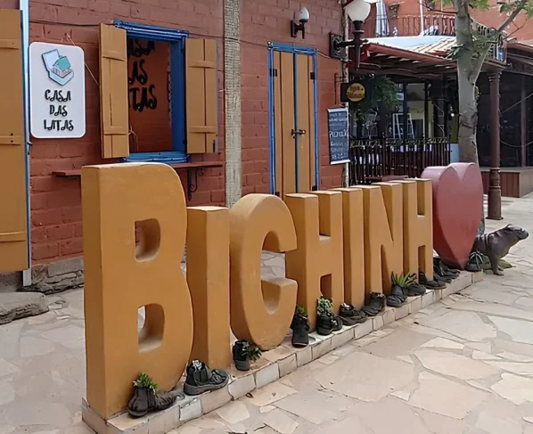 Escultura grande com as letras "BICHINHO" em um tom de amarelo-ocre, posicionada em uma calçada de pedra em frente à loja "Casa das Latas", com sapatos antigos servindo como vasos de plantas ao pé das letras e um coração vermelho ao lado.
